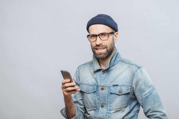 Atractivo modelo masculino de mediana edad con cerdas vestidas con ropa de moda, tiene teléfono inteligente