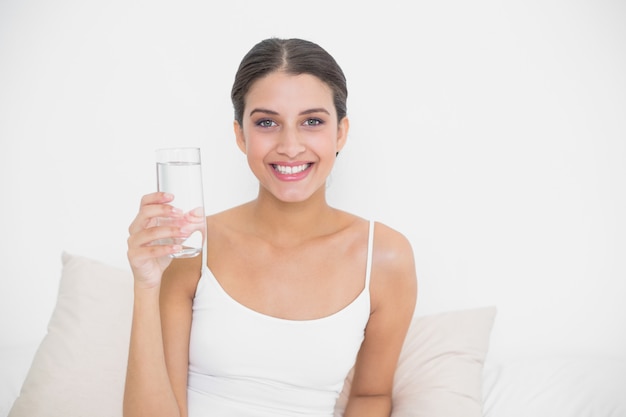 Atractivo modelo de pelo marrón joven en pijama blanco sosteniendo un vaso de agua