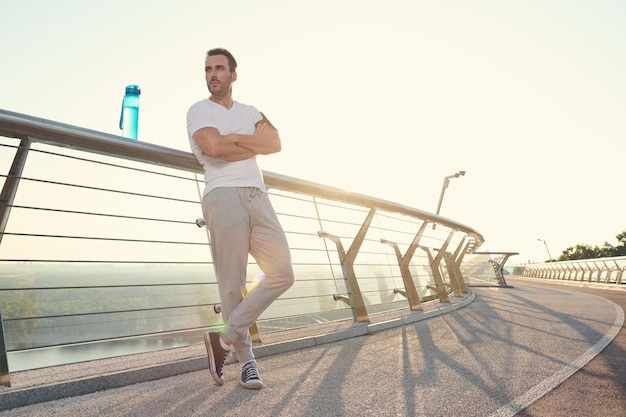 Atractivo musculoso de mediana edad, hombre caucásico, atleta en forma con un hermoso cuerpo estético que se relaja después de un fuerte entrenamiento al aire libre al amanecer, de pie en un puente de la ciudad de cristal