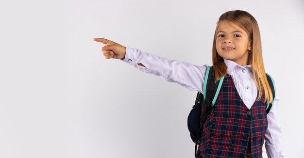 Atractivo niño rubio sonriente con cabello largo apuntando con el dedo índice para copiar el espacio en la pared blanca en blanco para su texto o anuncio.