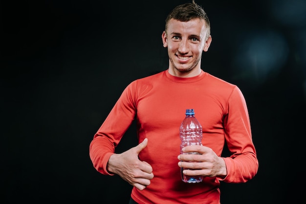 Atractivo retrato de un apuesto hombre caucásico musculoso sonriente que usa ropa deportiva roja con una botella de agua y pulgares hacia arriba después de un intenso entrenamiento físico en el gimnasio Motivación deportiva Estilo de vida