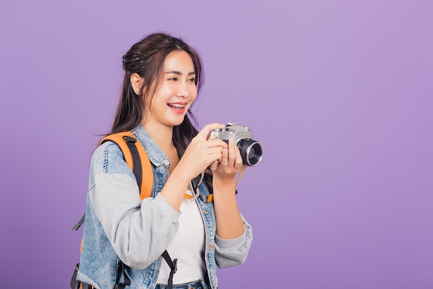 Atractivo retrato feliz asiática hermosa mujer joven sonriente emocionada usar jeans y bolso con cámara de fotos vintage, mujer viajera fotógrafa, foto de estudio aislada en fondo púrpura