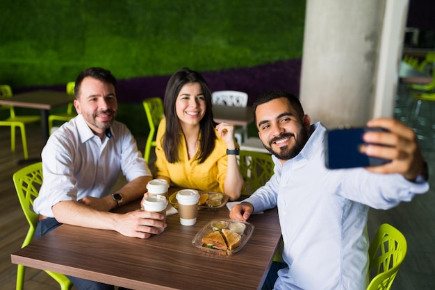 Atractivos amigos y compañeros de trabajo tomándose una selfie con un smartphone mientras se sientan juntos en una mesa en el patio de comidas y almuerzan después del trabajo