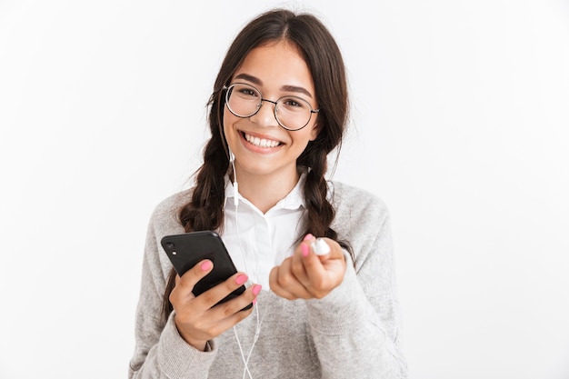 Atraente alegre colegial usando uniforme de pé isolado sobre uma parede branca, ouvindo música com fones de ouvido, segurando um telefone celular