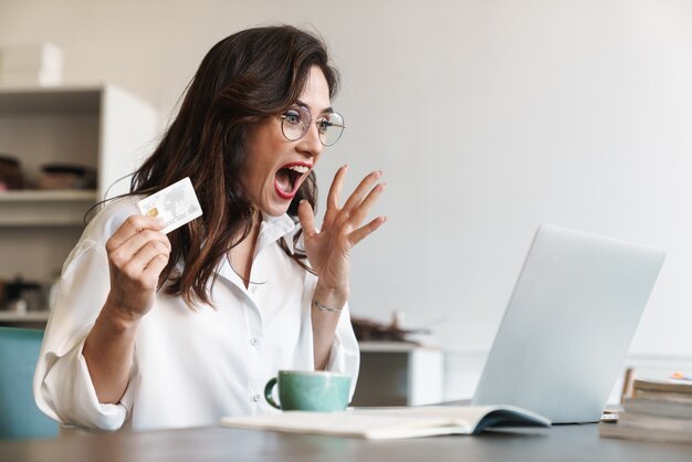 Atraente alegre jovem morena empresária sentada à mesa do café com o laptop dentro de casa, fazendo compras online com cartão de crédito