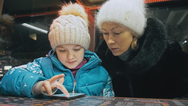 Atraente caucasiana mãe e filha usando smartphone sentado no café de inverno com chapéu de jaqueta Menina avançada mostra e explica a mãe mulher no smartphone tocando e assistindo