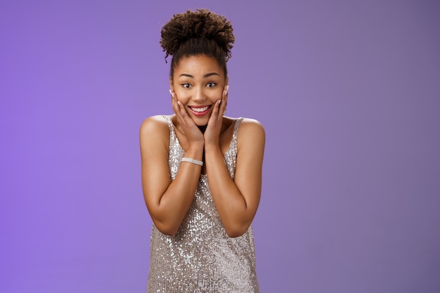 Atraente divertida encantadora mulher afro-americana fascinada em um elegante vestido prateado pressione as mãos as bochechas surpreso grato receba impressionante presente emocionante sorrindo encantado, fundo azul.