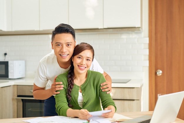 Atraente dona de casa asiática com expressão de felicidade enquanto calcula contas na cozinha