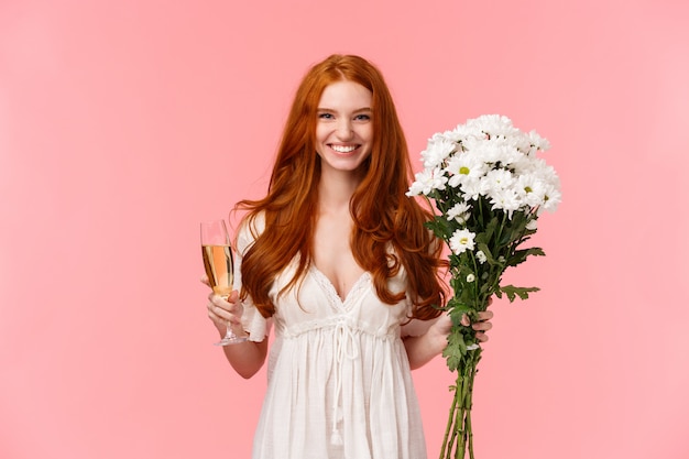 Atraente e animada namorada ruiva romântica recebe presente, segurando flores buquê branco e taça de champanhe, bebendo comemorando a ocasião, tendo o dia dos namorados perfeito, rosa