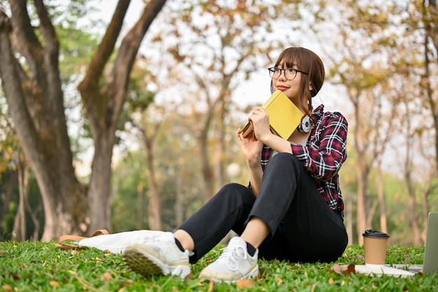 Atraente estudante universitária asiática senta-se na grama em um parque verde com seu livro