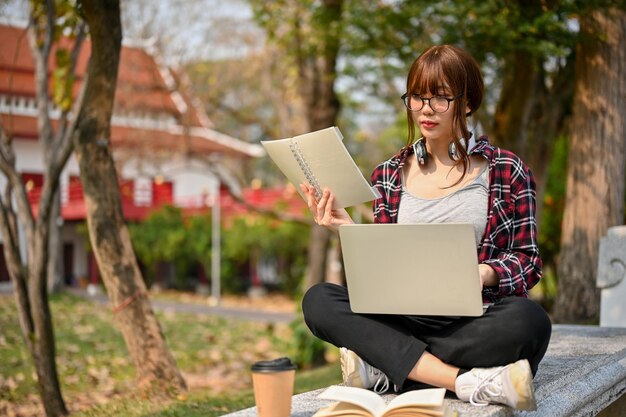 Atraente estudante universitária asiática usando laptop em um banco no parque do campus