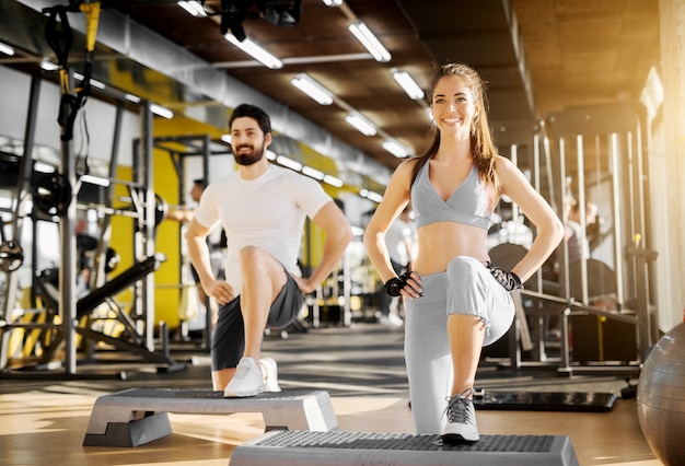 Atraente forte treinador muscular mostrando exercício de perna com stepper para uma linda garota sorridente com luvas no ginásio.