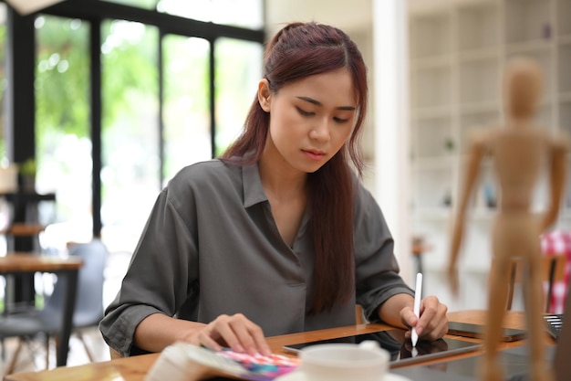 Atraente jovem designer gráfico feminino asiático senta-se em sua mesa de escritório usando tablet gráfico