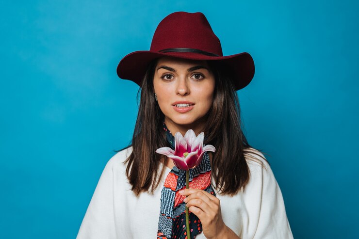 Foto atraente jovem detém flor, olhando apaixonado, vestido de suéter branco, chapéu cor de vinho com uma aba e lenço colorido no pescoço, sorri timidamente. isolado sobre o fundo azul