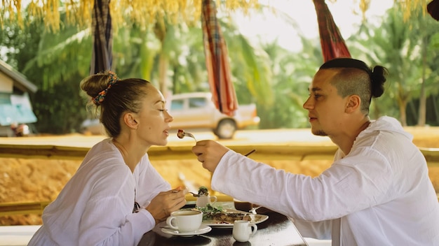 Atraente jovem e mulher comem e bebem bebidas saborosas, passando o tempo no tradicional café flutuante local na água Amando casal feliz tomando café da manhã ao ar livre se alimentam vista tropical