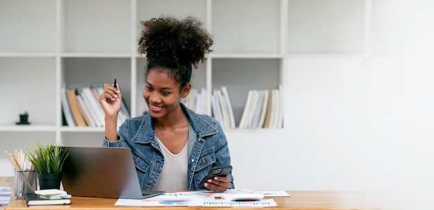 Atraente jovem empresária africana confiante sentada à mesa do escritório trabalhando em laptop e telefone celular