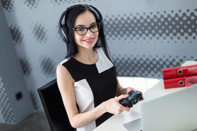 Atraente jovem empresária de vestido preto, fones de ouvido e óculos sentar à mesa e jogar no laptop