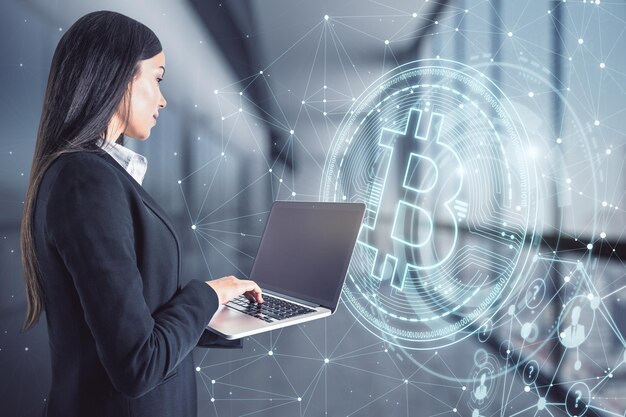 Atraente jovem empresária europeia usando laptop no interior do escritório embaçado com holograma de bitcoin brilhante Criptomoeda e conceito de finanças Dupla exposição