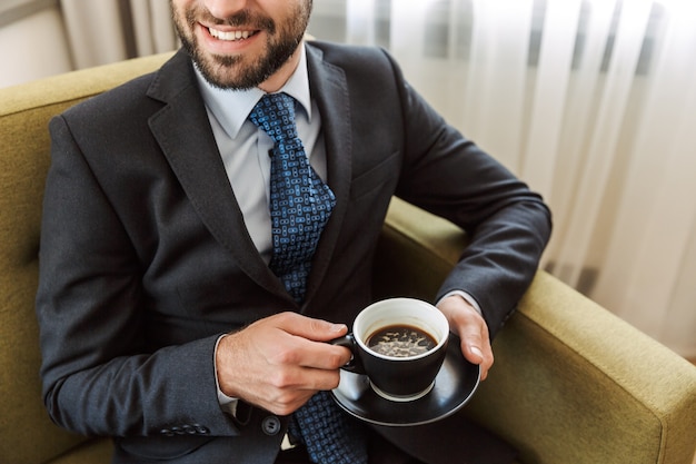 Atraente jovem empresário vestindo terno, sentado em uma cadeira no quarto do hotel, segurando uma xícara de café