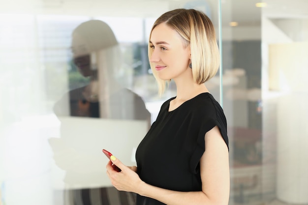 Atraente jovem loira empresária em uma elegante blusa preta com smartphone no escritório da empresa