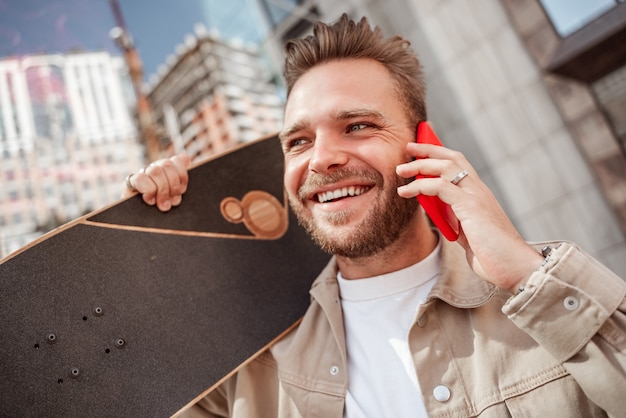 Atraente jovem loira sorridente segurando o skate nos ombros em pé de rua da cidade entre edifícios de vidro. Usa roupa jeans. Skatista estiloso e esportivo fala sobre lauphing de celular.