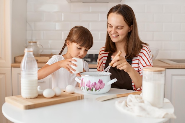 Atraente jovem mãe satisfeita e sua filha assando na cozinha em casa preparando pastelaria caseira juntos expressando felicidade criança do sexo feminino anúncio açúcar