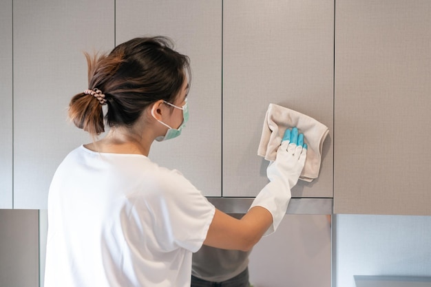 Atraente jovem mulher asiática usando luvas brancas protetoras de borracha limpando uma superfície do armário de parede da cozinha com pano Conceito de limpeza doméstica