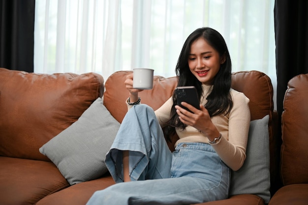 Atraente jovem mulher asiática usando seu smartphone enquanto está sentada e tomando café no sofá