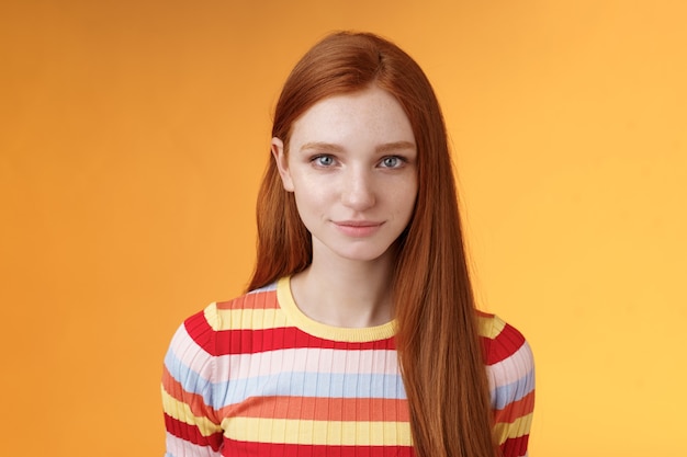 Atraente jovem ruiva sincera limpa puro perfeito tom de pele sorrindo modesto olhar câmera amigável encantado em pé relaxado esperando olhando boba ternamente, posando de fundo laranja. Copie o espaço