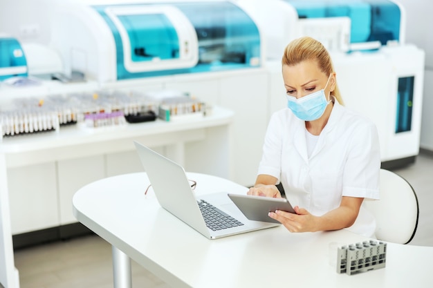 Atraente loira assistente de laboratório vestindo jaleco com máscara cirúrgica estéril sentado no laboratório