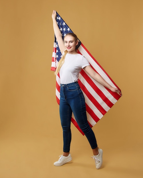 Atraente loira posa com orgulho com uma bandeira americana. sessão de fotos no estúdio em um fundo amarelo.