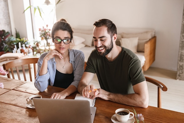 Atraente morena casal homem e mulher bebendo café e trabalhando juntos no laptop enquanto estão sentados à mesa em casa