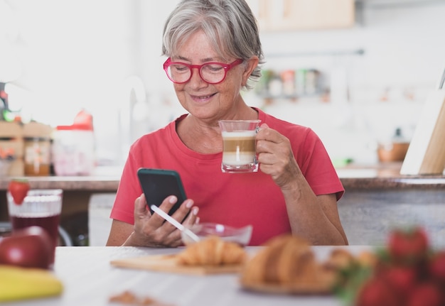 Atraente mulher aposentada sênior, desfrutando do café da manhã em casa com croissant e cappuccino. Olhando as notícias no celular