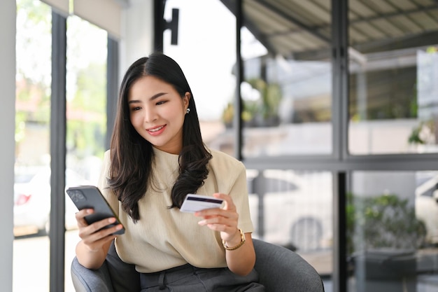 Atraente mulher asiática segurando seu smartphone e cartão de crédito usando o aplicativo bancário móvel