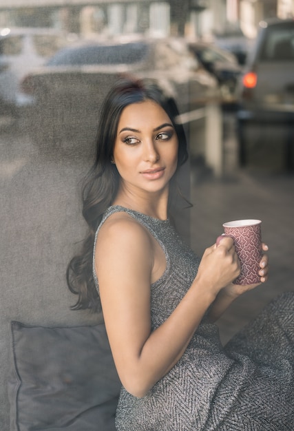 Foto atraente mulher indiana segurando a xícara de café, olhando pela janela, sentado no café
