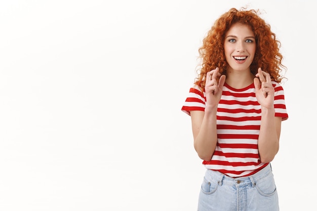 Atraente otimista alegre sorridente ruiva cacheada mulher olhos azuis cruzam os dedos boa sorte sorrindo esperançosamente antecipando boas notícias em pé parede branca