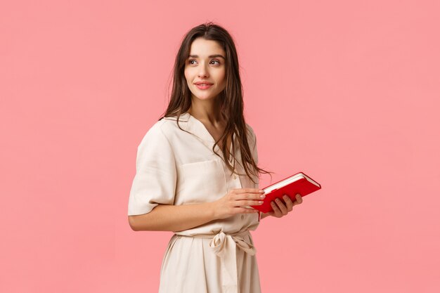 Foto atraente sonhadora e romântica linda garota de vestido vire pensativo, segurando o caderno, prepare notas para o trabalho, parede rosa de pé, pensando