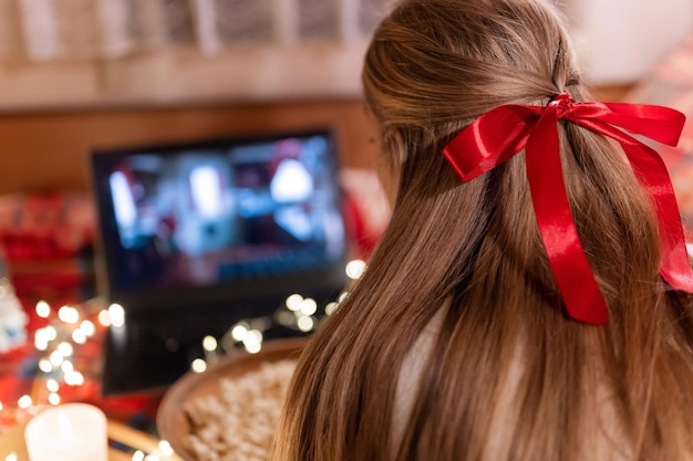 Atrás da cabeça feminina com longos cabelos loiros e vista traseira do laço vermelho assistindo o filme de natal no laptop no acampamento em casa na cama aconchegante com guirlanda de luzes brilhantes à noite na véspera de natal ou ano novo