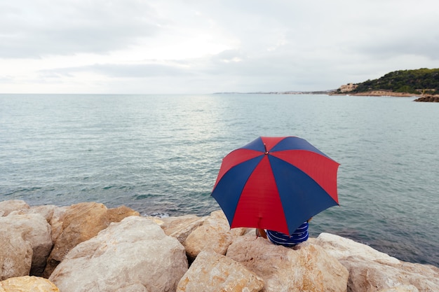 Desde atrás, un hombre anónimo sentado en las rocas con un paraguas junto al mar mientras llueve.