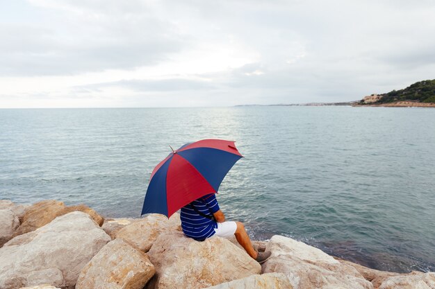 Desde atrás, un hombre anónimo sentado en las rocas con un paraguas junto al mar mientras llueve.