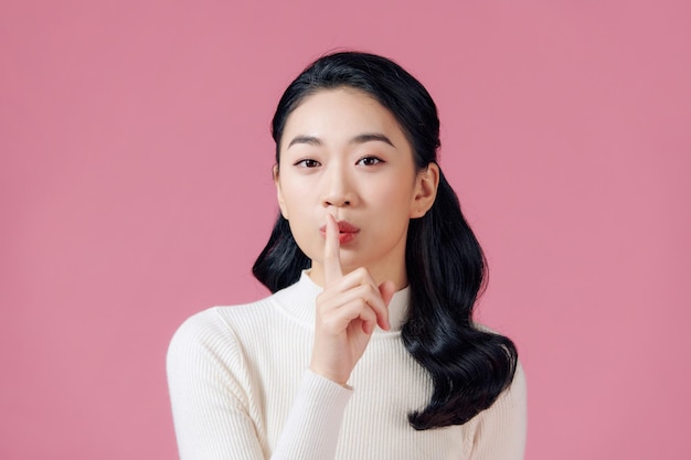 Atrevida y creativa linda chica asiática callándose con un dedo sobre el labio sonriendo misteriosa o astuta