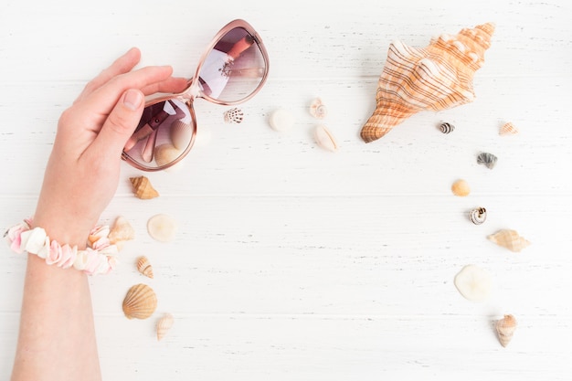 Atributos de las vacaciones de verano. Gafas de sol rosadas en mano femenina con diferentes conchas