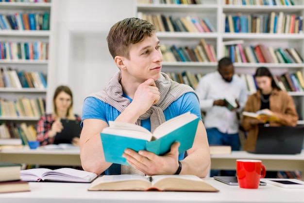 Attraktiv denkender hübscher männlicher Student, der mit vielen Büchern am Tisch sitzt und sich auf Test oder Prüfung vorbereitet