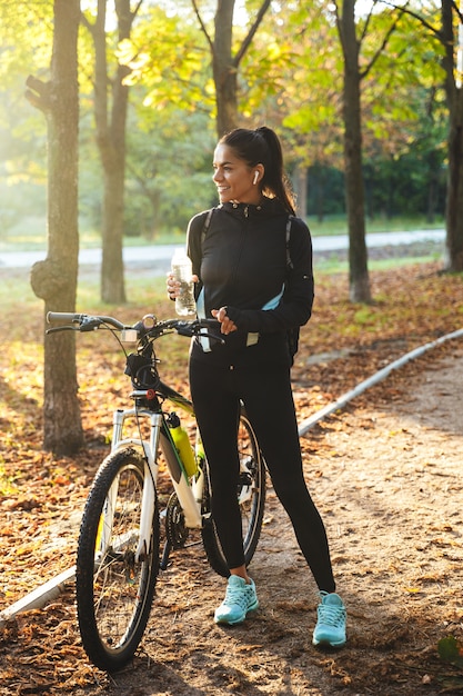 Attraktiv fit Sportlerin mit Fahrrad im Park, Trinkwasser aus einer Flasche