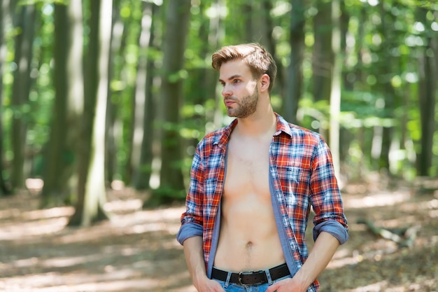 Attraktiv und charismatisch. Sexy Mann steht im Holz auf Naturlandschaft. Schöner Kerl mit sexy nacktem Oberkörper im offenen Hemd. Sommerurlaub. Fernweh und Reisen.