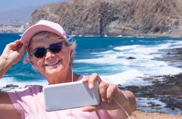 Attraktive ältere Frau, die Handy hält, das an windigen Tagen selfie auf See nimmt. Senioren genießen Urlaub und Schönheit in Natur, Bergen, Strand und Wellen im Hintergrund