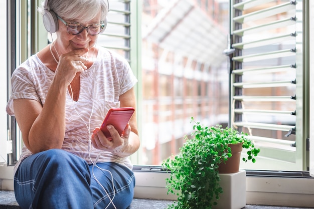 Attraktive ältere Frau, die nahe am Fenster mit tragenden Kopfhörern des Mobiltelefons sitzt. Lächelnde kaukasische Frau, die den Lebensstil im Ruhestand genießt