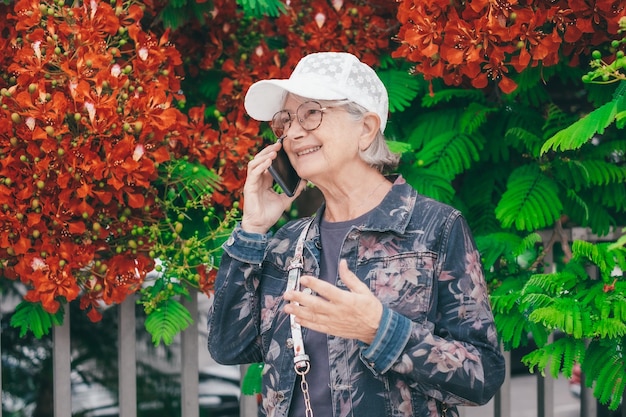 Attraktive ältere Frau in Jacke-Jeans und weißer Mütze geht im Frühlingspark im Freien mit dem Telefon