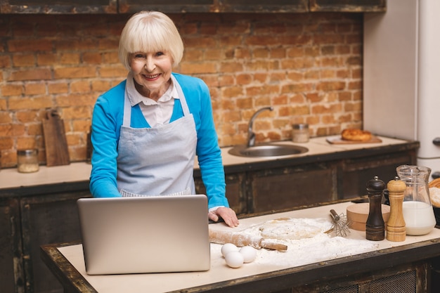 Attraktive ältere Frau kocht auf Küche. Großmutter macht leckeres Backen. Laptop benutzen.