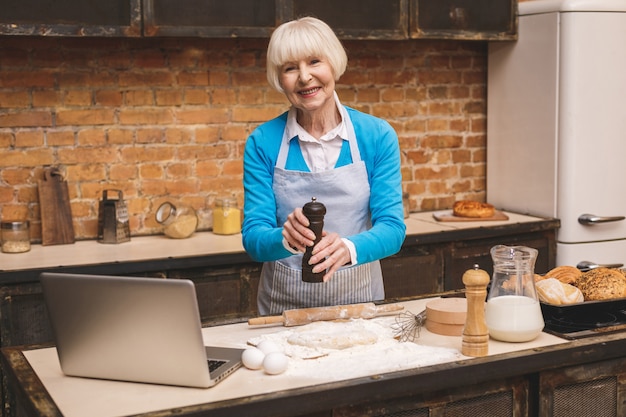 Attraktive ältere Frau kocht auf Küche. Großmutter macht leckeres Backen. Laptop benutzen.